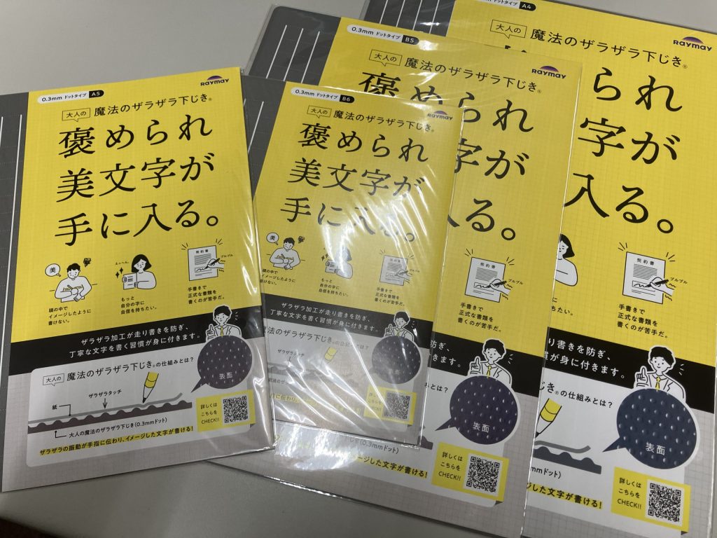 この画像は「魔法のザラザラ下じき」のパッケージが並んでいる様子です。黄色いパッケージには、「擦（す）れられ美文字が手に入る。」というキャッチフレーズが書かれており、筆記時に滑りにくくすることで、綺麗な文字を書くサポートをすることが強調されています。製品の特徴として、書き心地を良くするために「ザラザラ加工」が施された下敷きが含まれており、さまざまなサイズのものが販売されていることが示されています。