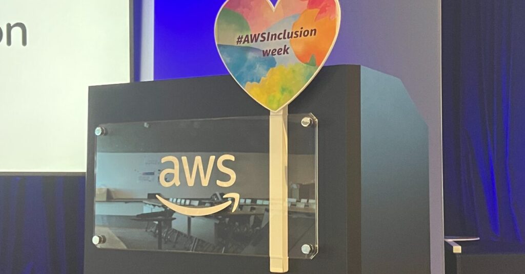 AWSロゴの演台の前に、「#AWSInclusion week」と書かれたカラフルなハート形のうちわが立てかけられている