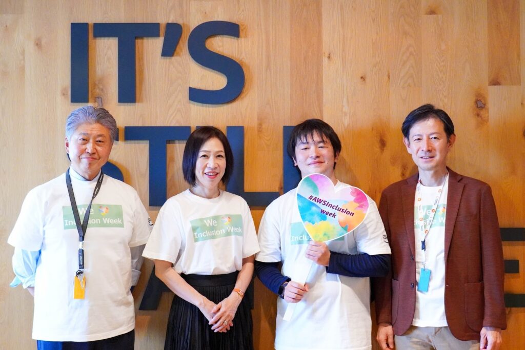 「IT’S STILL」と書かれた木目の壁の前で4人が並んで記念撮影。1人が「#AWSInclusion week」のハート形うちわを持ち、全員が「We Inclusion Week」Tシャツを着ている。
