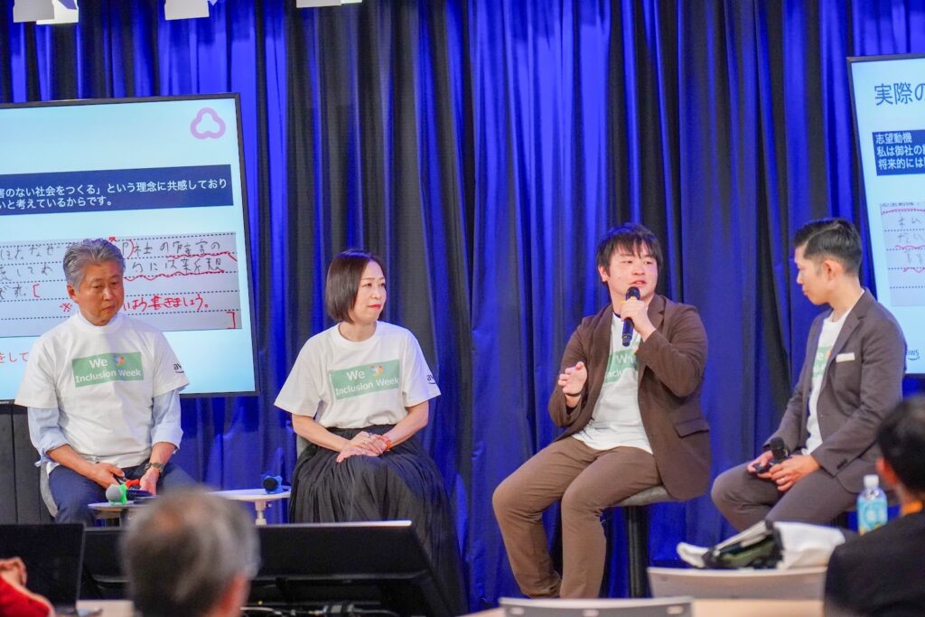 青いカーテンの前のステージで4人が着席してパネルディスカッション。中央の男性がマイクで発言し、他の3人が聞いている。